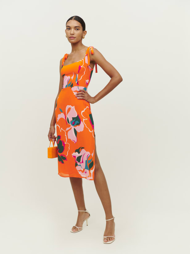 Reformation Besse dress is a midi length dress with a scoop neckline. It features tie straps and back smocking for a little extra bit of stretch. It has a high, side slit to show off your figure and allow movement and it comes in an orange-based print with oversized tropical looking flowers. This would make a stunning wedding guest or holiday dress. 
