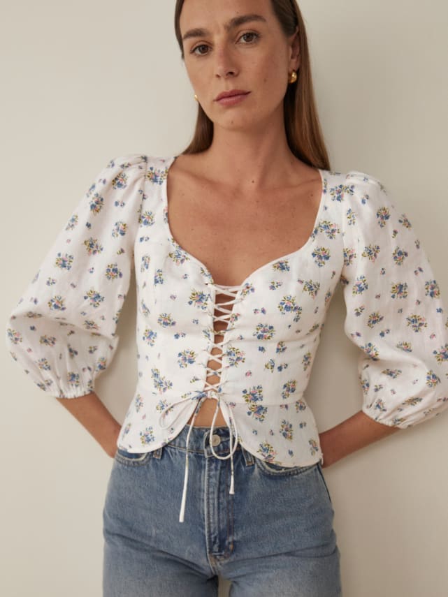 3/4 length sleeve top with an adjustable, lace up front and a fitted bodice. It features a sweetheart neckline and voluminous sleeves. White linen with ditsy floral print. 
