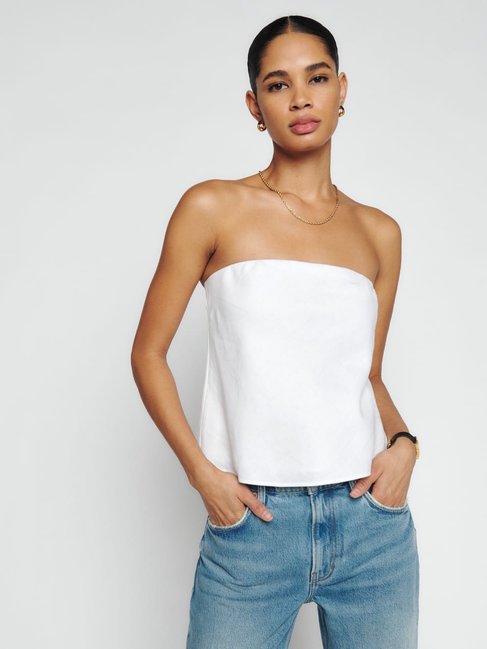 Reformation spritz linen top in white. It has an elastic bandeau neckline. Non-adjustable straps. Lightweight 100% linen fabric. linen. The model is wearing it with blue jeans. 